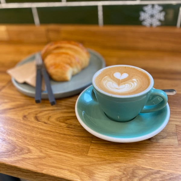 Turquoise cup of cappuccino with latte art on a wooden table, accompanied by a croissant.