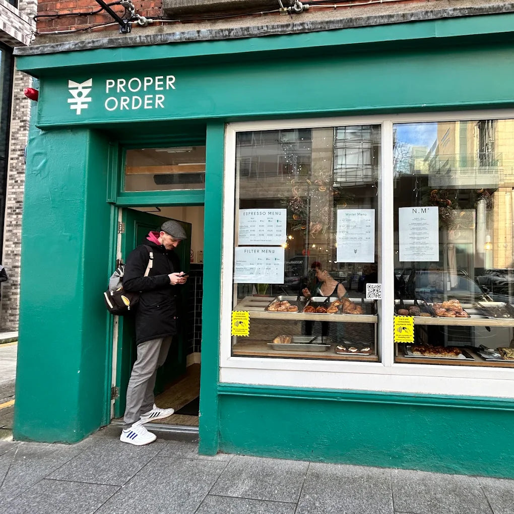 Green storefront of 'Proper Order Coffee Company' with people inside and pastries displayed.