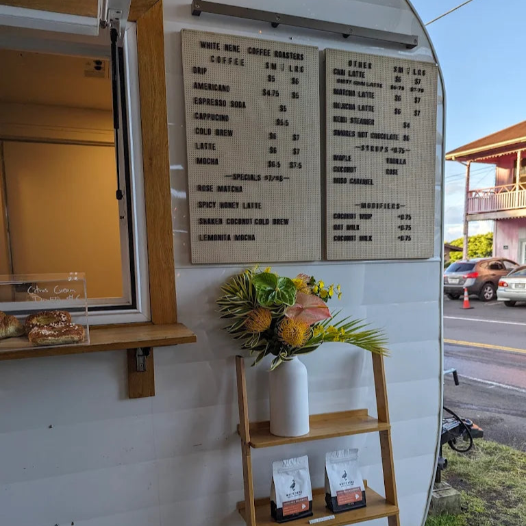 Third wave coffee truck with menu board, flowers, and coffee bags on a wooden shelf in Kona Hawaii.