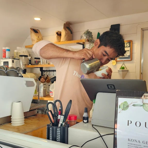 Person working in a specialty coffee shop pouring a drink, surrounded by coffee-making equipment and decor in Hawaii.